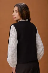 Chaleco de punto negro con cuello redondo y corte recto. Presenta aberturas laterales amplias con detalle de tira y botón, ofreciendo un diseño versátil y moderno para superposiciones.