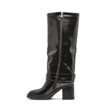 Bota de caña alta Korium modelo Sira, confeccionada en cuero sintético negro con acabado brillante. Presenta un taco cuadrado robusto, punta redondeada y una tira decorativa con hebilla metálica en la zona del tobillo.