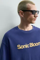 Remera de algodón con cuello redondo y manga corta, de corte holgado. Presenta un estampado frontal con la frase 'Sonic Bloom' en color amarillo.