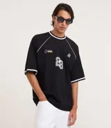 Remera deportiva de algodón, color negro, con corte oversized, cuello redondo y mangas cortas. Presenta ribetes contrastantes en blanco en el cuello y los bordes de las mangas. Tiene un estampado localizado en el pecho con tipografía en relieve y un pequeño logo en el lateral superior.