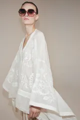 Blusa tipo capa de algodón con escote en pico y cierre frontal con botones. Presenta mangas largas amplias y diseño con bordados florales a tono, con bajo asimétrico.