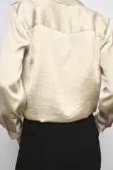 Camisa de cuello solapa y manga larga con puños anchos, confeccionada en tejido satinado color beige. Presenta cierre frontal con botones ocultos y bajo ajustable mediante cordones del mismo tejido.