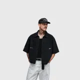 Camisa de jean negra de manga corta, con cuello clásico, cierre frontal de botones y bolsillos frontales tipo parche. Presenta detalles de estampado minimalista en el pecho y la manga.