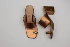 Sandalias de cuero color bronce con taco cuadrado y tira ancha sobre el empeine. Presentan un aro metálico plateado que separa el dedo gordo del resto.