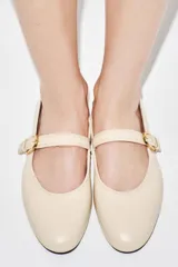 Zapato plano tipo bailarina confeccionado en piel de color crudo, con tira en el empeine y hebilla metálica dorada ajustable. Presenta puntera redonda y suela plana.
