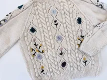 Sweater de lana merino color beige, con tejido de punto trenzado y bordados artesanales de flores en tonos azules, amarillos y morados, cuello redondo y mangas largas.