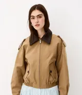 Campera Comfort de poliéster color camel, con cuello de poliuretano en contraste color marrón oscuro, cierre frontal y bolsillos con solapa y botones. Presenta martingalas en los hombros y elástico en la cintura.