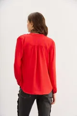 Camisa roja de manga larga con cuello redondo y escote frontal con botones ocultos. Presenta detalles de alforzas y frunces en los hombros.