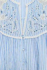 Blusa de rayas verticales blancas y celestes, con cuello redondo y escote en pico. Presenta detalles de bordados perforados y puntillas en el pecho y las mangas. Tiene mangas largas abullonadas con puños elásticos y cierre frontal con botones forrados.