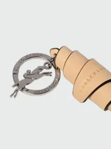 Llavero Longchamp de la línea Autres Lignes, modelo Croissant, confeccionado en cuero color tostado con detalles grabados de la marca y herrajes metálicos plateados con el logo distintivo de un jinete a caballo.