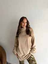 Buzo de peluche color beige, con cuello alto y diseño holgado. Presenta un parche cuadrado con logo en la manga derecha.