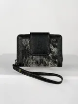 Billetera pequeña tipo clutch con correa de muñeca, confeccionada en cuero negro texturizado y un panel central de pelo (pellejo) en tonos blanco y negro. Presenta costuras blancas de contraste y un cierre de solapa con broche.