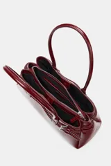 Bolso de hombro tipo city en color bordó con acabado brillante. Presenta un diseño estructurado con detalle de tiras decorativas con hebillas metálicas en los laterales y doble asa de hombro.