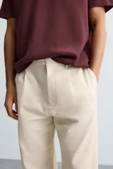 Pantalón de corte recto, color beige claro, con pliegues frontales y bolsillos de vivo en la parte trasera.