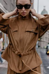 Camisa de estilo safari en color marrón con efecto ante, cuello subido y mangas anchas de largo tres cuartos con terminación en vuelta. Presenta bolsillos con solapa en el pecho, cintura ajustable con lazo del mismo tejido y cierre frontal oculto con cremallera y botones a presión.