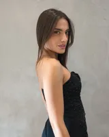 Vestido corto strapless negro con diseño drapeado en toda la prenda, detalle de aplique floral en el centro del escote y caída en cascada con volados frontales. Presenta pequeños puntos brillantes distribuidos por toda la superficie.