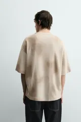 Camiseta de punto de algodón con corte relajado (relaxed fit) y cuello redondo. Presenta un estampado frontal con efecto spray en tonos marrones y beige, y un efecto lavado general.