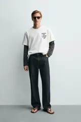 Camiseta blanca de corte relajado, con cuello redondo y manga corta. Presenta un pequeño estampado de texto en contraste en la parte frontal izquierda del pecho.