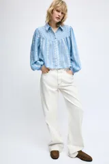 Blusa corta de color blanco con diseño de bordados perforados, cuello solapa, cierre frontal con botones y mangas largas abullonadas.