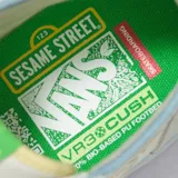 Zapatillas de caña alta Vans x Sesame Street, color blanco con detalles en rosa, verde y celeste. Presentan un dibujo de Elmo en el lateral y un dije de felpa con la letra L.