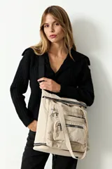 Mochila shopper color beige, confeccionada en poliéster. Presenta un diseño versátil con múltiples cierres, compartimento inferior expandible y un bolsillo frontal removible que funciona como estuche. Incluye correas ajustables y removibles para usar como mochila o bandolera, además de asas fijas para llevar al hombro.