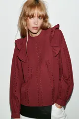 Blusa de manga larga con cuello redondo, volantes en los hombros y detalle de jaretas frontales. Presenta bordados perforados en toda la prenda y cierre posterior con botones ocultos.