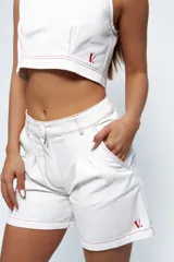 Short de gabardina color blanco con costuras a la vista en color rojo. Presenta un diseño de tiro medio-alto con tablones delanteros, bolsillos laterales y traseros, y cierre frontal con botón. Incluye un pequeño bordado con la letra V en color rojo en la parte inferior.