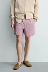 Bermuda de hombre con corte relaxed fit, tiro medio, cintura elástica con botón y cierre de cremallera. Presenta un patrón de cuadros pequeños en tonos rosados y grises.