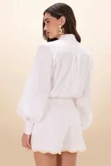 Camisa de tricolina color blanco, con cuello clásico, mangas largas abullonadas y puños ajustados. Presenta detalles de bordado calado con motivos florales en la parte superior delantera.