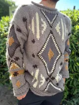 Sweater de lana tejido a mano con diseño étnico, corte oversize y mangas amplias. Presenta un patrón geométrico con rombos y estrellas en tonos tierra sobre una base gris.