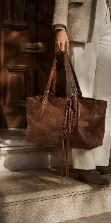 Cartera tipo tote de cuero vacuno color marrón, con diseño artesanal, asas reforzadas y detalle de flecos laterales largos.