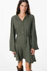 Vestido corto de color verde militar, con cuello camisero, cierre frontal de botones y mangas largas con puños con volados. Incluye un lazo ajustable en la cintura y falda con vuelo.