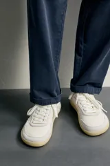 Zapatilla deportiva barefoot fabricada en piel, en color blanco con detalles en beige y gris, y suela de goma en contraste.
