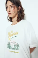 Remera blanca de manga corta con calce relajado y cuello a la base. Presenta un estampado frontal pequeño y una estampa protagonista en la espalda con el texto "Rocky Mountain National Park" y un paisaje de montañas y árboles.