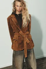 Blazer de gamuza color marrón con cuello solapa, manga larga con hombreras y cierre frontal con botones. Incluye bolsillos con solapa en el delantero, bolsillo de vivo en el pecho y cinturón de lazada en el mismo material.