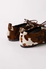 Zapato tipo flat de estilo acordonado, confeccionado en cuero con pelo con estampado de manchas tipo vaca en tonos marrón y blanco.