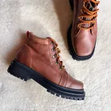 Botas de cuero color marrón con suela track negra gruesa, cordones naranjas y cierre frontal con cremallera.