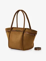 Bolso tipo shopper de eco suede color marrón, con forma trapezoidal y asas de hombro. Presenta detalles de cuero marrón con pespuntes blancos y un broche metálico dorado en el centro superior. Incluye correa larga desmontable.