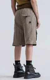 Bermuda técnica color arcilla (marrón claro/beige) de calce oversize, confeccionada en poliéster liviano. Presenta múltiples bolsillos cargo con tapa y un cinturón de ajuste con hebilla plástica negra. Incluye detalles reflectivos.