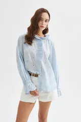 Camisa de algodón con detalles de broderie, volados y cuello a la base. Presenta diseño holgado, cierre frontal con botones y mangas largas con puños fruncidos.