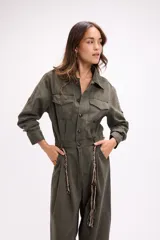 Mono de estilo utilitario color beige, con cierre frontal de botones, bolsillos en el pecho y cintura entallada con detalle de cadenas decorativas. Presenta mangas largas y piernas de corte recto y holgado.