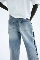 Jean de tiro alto, confeccionado en denim de algodón rígido, de corte ancho y pierna recta. Presenta un lavado celeste claro con efecto desgastado en el centro y franjas laterales en denim más oscuro con acabado desflecado.