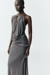 Vestido largo de color negro con cuello halter y detalle de aro metálico en el escote. Presenta un diseño drapeado en la parte frontal y escote en pico pronunciado.