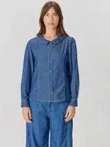 Camisa de denim con silueta levemente entallada, cuello bebé y mangas largas con sutil frunce en los hombros. Presenta costuras curvas decorativas con pespuntes en contraste y cierre frontal con botones.
