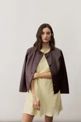 Chaqueta corta de cuero color bordó, de diseño estructurado, sin cuello, con cierre frontal de botones y detalles de costuras decorativas.