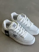 Championes DC modelo Manteca, color blanco con detalles en gris, cordones anchos y suela de goma marrón. Presentan el logo de DC en la lengüeta y los laterales.