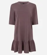 Vestido tipo camiseta de algodón, color malva o ciruela con efecto lavado, de corte recto y holgado, con cuello redondo y mangas cortas. Presenta un volado fruncido en el bajo.