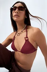 Top de bikini color borgoña, de diseño triangular y textura arrugada. Se ata en el cuello y la espalda con tiras finas. Los tirantes tienen apliques de aros cuadrados.