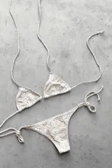 Conjunto de bikini con corpiño triangular de atar con apliques de argollitas y bombacha vedetina de atar con apliques de argollitas. El tejido presenta un estampado tie-dye o marmolado en tonos lila/azul y naranja/beige.