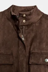 Camisa de efecto ante en color marrón, con cuello subido, mangas tres cuartos con vuelta y cierre frontal con cremallera. Presenta falsos bolsillos con solapa en el pecho y cintura ajustable con cordones del mismo tejido.